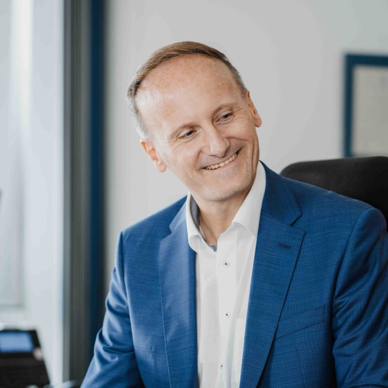 Mann mit freundlichem Lächeln in den Kanzleiräumen in Großwallstadt (Landkreis Miltenberg), trägt einen blauen Anzug und ein weißes Hemd.