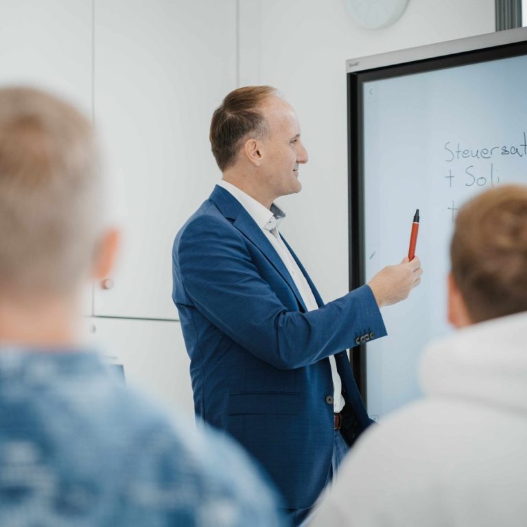 Mann steht vor einer digitalen Tafel in den Kanzleiräumen in Großwallstadt und erklärt einer Gruppe von Schülern Lohnabrechnung.
