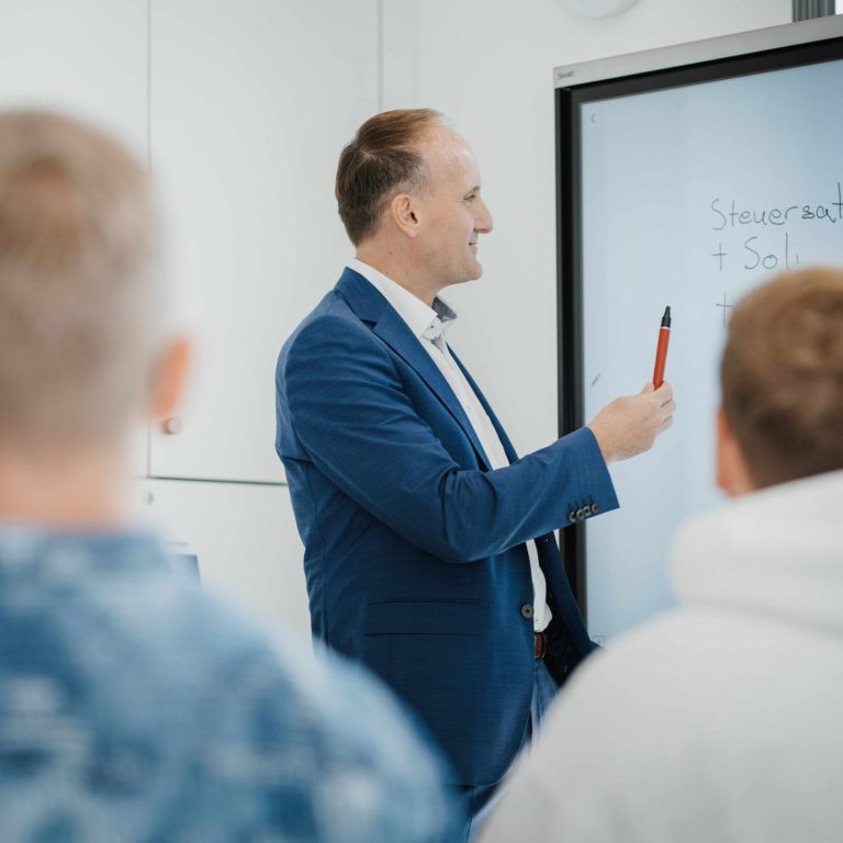 Mann steht vor einer digitalen Tafel in den Kanzleiräumen in Großwallstadt und erklärt einer Gruppe von Schülern Lohnabrechnung.